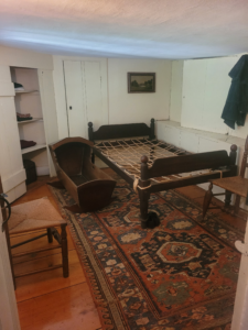 A small room with a low ceiling. The contents of the room include a bed, cradle, chairs, rug, closet, and small painting. 