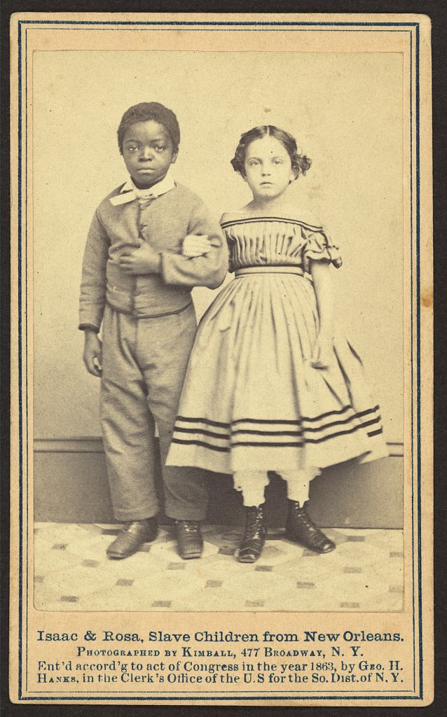 Two enslaved children posing for a photograph. A young boy on the left has dark skin and is dressed in a simple suit. The girl on the right has very light skin and is wearing a dress. Both are looking directly at the camera with a serious expression and are standing in front of a plain wall.