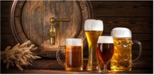 Four differently-shaped glasses, all filled with beer. In the background are a keg and sheaf of wheat.