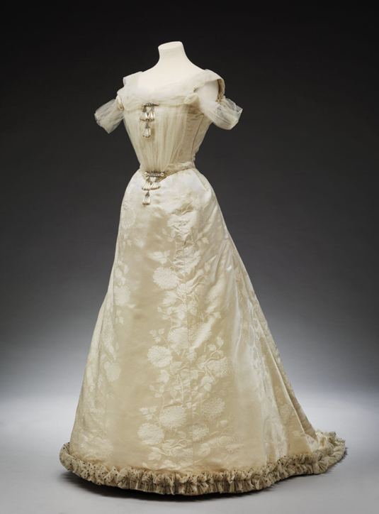 A floor length white cream gown, tulle on the bust and shoulders, decorations along bottom hem.